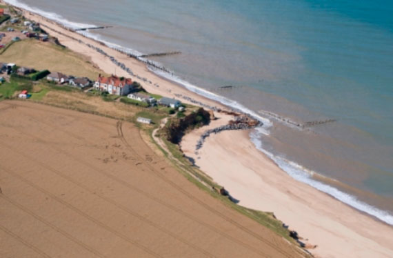 Aerial view of cliff erosion at Happisburgh on the East Coast of Norfolk. The end of Beach Road and the properties there on perch perilously close to the cliff edge, protected by stone escarpments on the sandy beach below and wooden spurs out into the blue sea water.